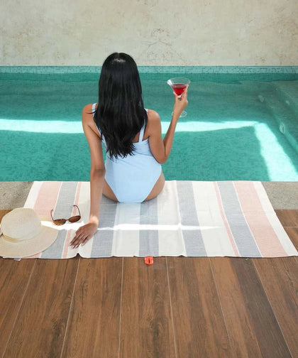 Woman in a light blue swimsuit sitting by a pool holding a drink, with a bamboo dobby towel and hat on the floor.