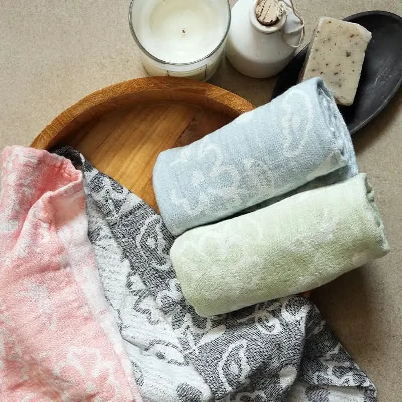 Set of folded towels in coral, charcoal grey , chalk blue and pastel green on a wooden tray with a candle and soap in the background.